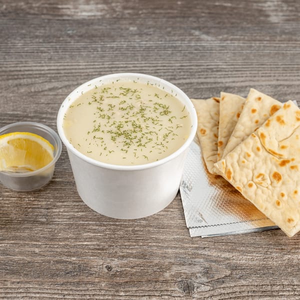 Creamy Greek soup with dill in a white cup, served with pita bread and a lemon wedge.